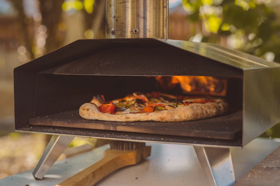 Four à pizza extérieur avec pizza en cuisson, flammes visibles à l'arrière, cheminée métallique, installation sur terrasse avec végétation en arrière-plan