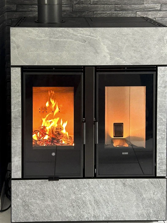 Une photographie en gros plan d'un poêle mixte moderne en acier ou en fonte, installé dans un salon, montrant clairement l'appareil avec son foyer visible permettant d'observer les flammes du bois et les granulés, dans un intérieur résidentiel contemporain.